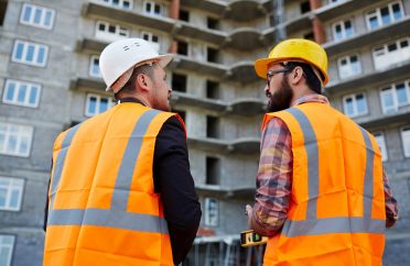 Backs of two builders in uniform having discussion on background of unfinished edifice